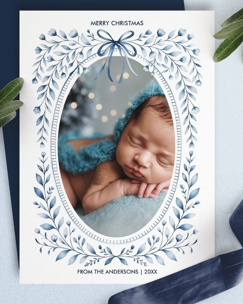 Christmas card featuring a sleeping baby in a blue bonnet, framed by a blue floral design.