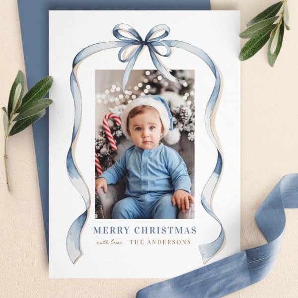 Christmas Card Featuring A Baby In A Blue Outfit And Santa Hat. A Blue Watercolor Ribbon Design Frames The Photo.