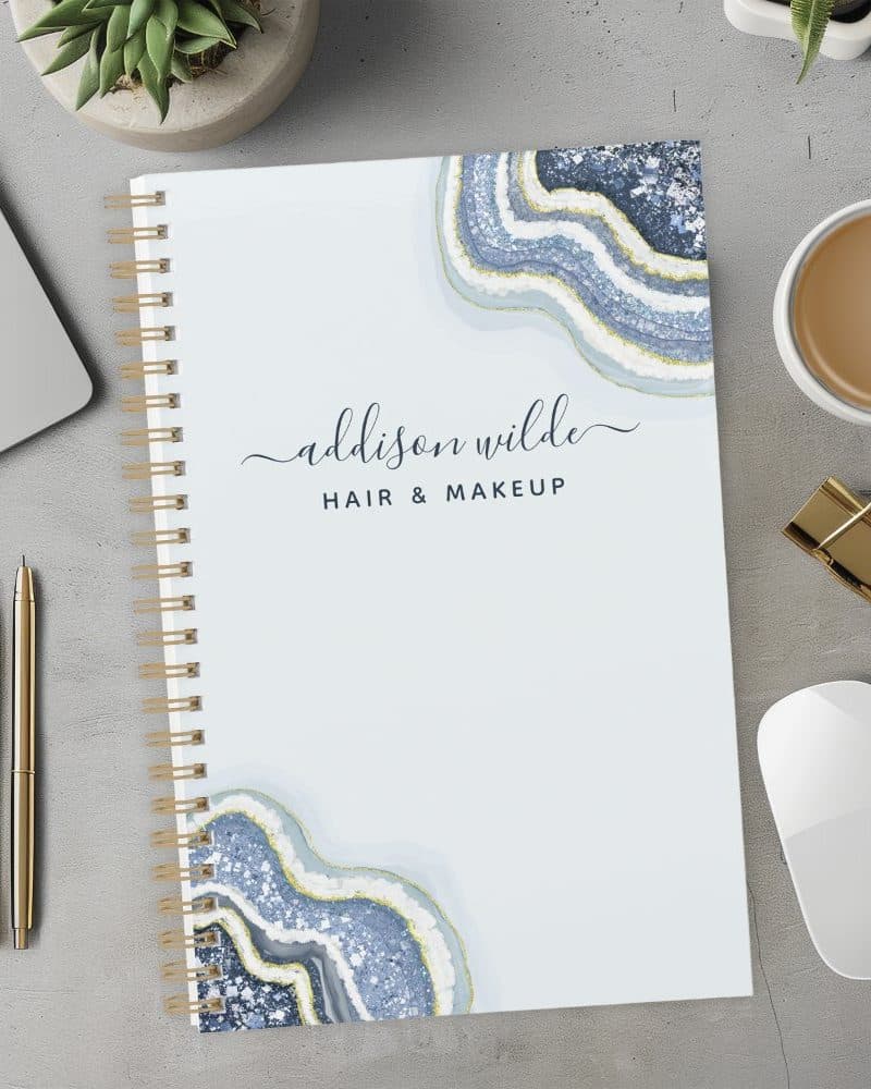 Spiral-bound notebook with a light blue cover featuring an agate design and the name Addison Wilde Hair & Makeup.