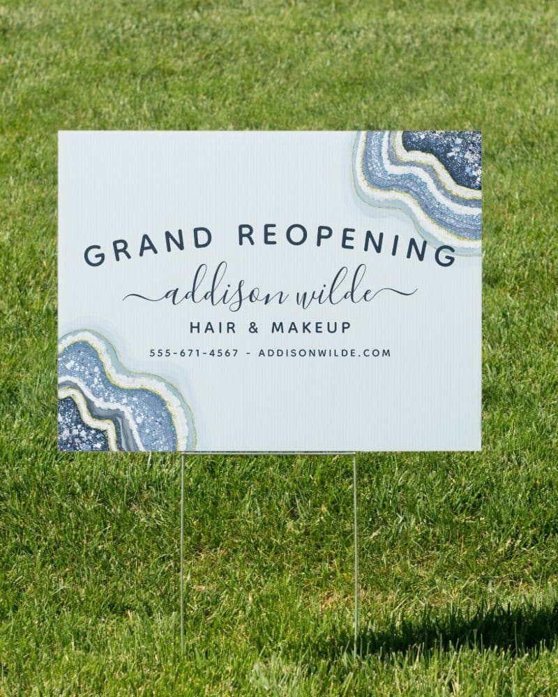 A yard sign announcing the grand reopening of Addison Wilde Hair & Makeup. The sign features a blue and gold agate design.