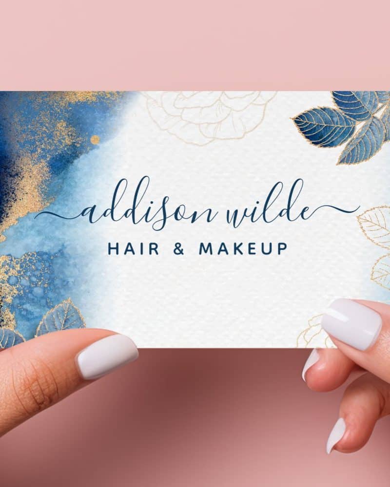 Two hands hold up a business card for Addison Wilde Hair & Makeup. The card features a blue and gold watercolor design with floral accents.