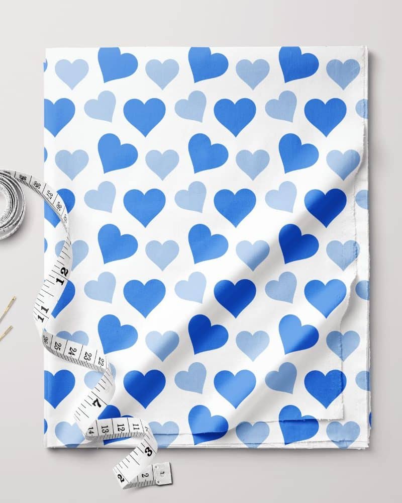 White fabric with a repeating pattern of light and dark blue hearts, accompanied by a measuring tape and sewing needles.