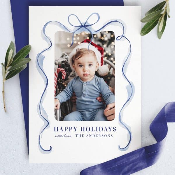 Christmas Holiday Card With A Photo Of A Baby In A Blue Outfit And Santa Hat, Framed By A Blue Watercolor Ribbon Design. The Andersons' Names Are Below.