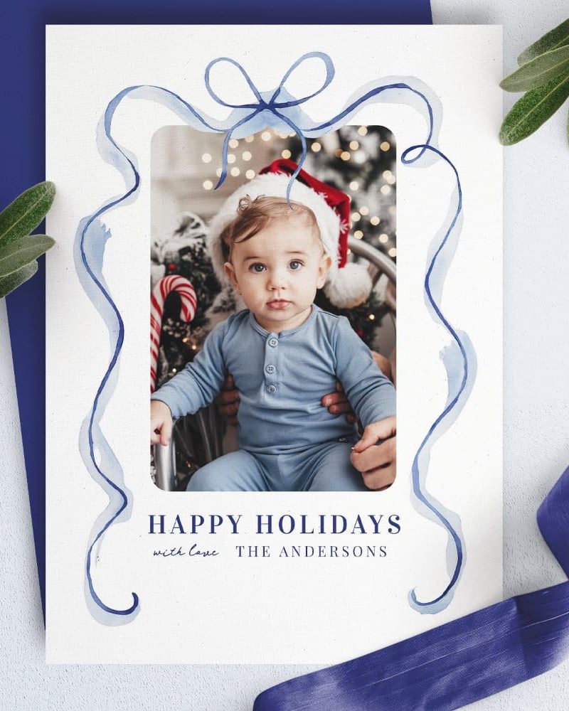 Christmas holiday card with a photo of a baby in a blue outfit and Santa hat, framed by a blue watercolor ribbon design. The Andersons' names are below.