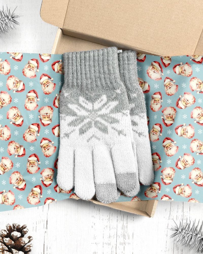 A pair of gray and white knit gloves with a snowflake pattern, nestled in a gift box lined with Santa Claus themed wrapping paper.