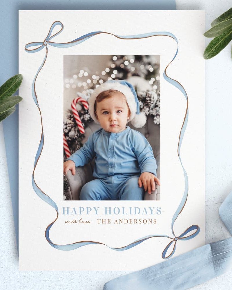 Christmas holiday card featuring a photo of a baby in a blue outfit and Santa hat. A blue ribbon border surrounds the image.