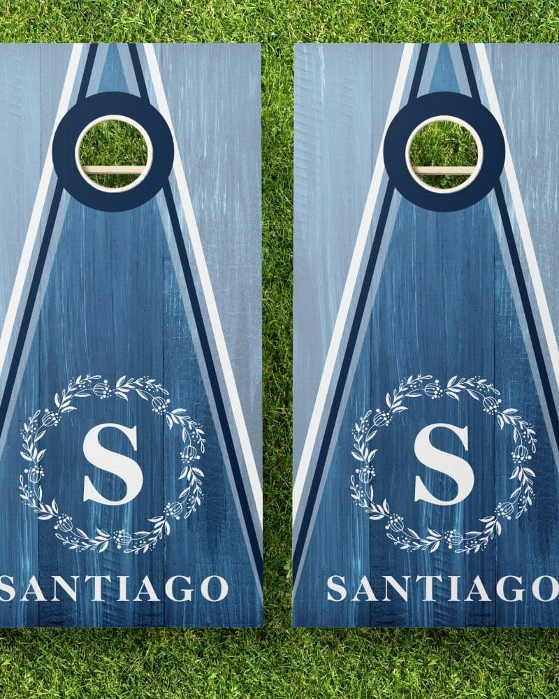 Pair of blue cornhole boards with white lettering. Each board displays a monogram in a wreath and the name Santiago.