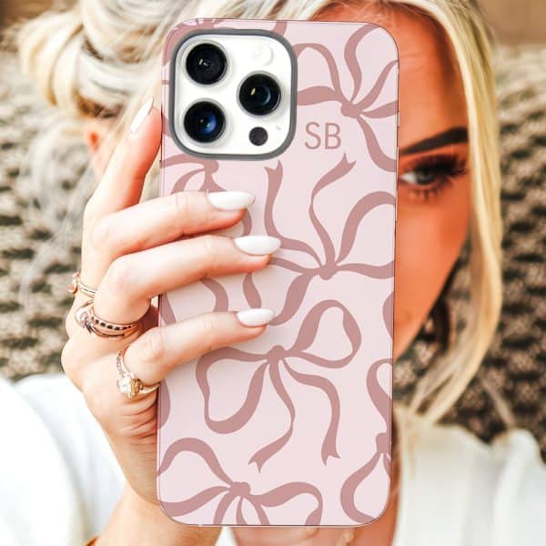A Woman Holds Up A Pink Iphone Case With A Pattern Of Mauve Bows. The Initials Sb Are Visible On The Case.