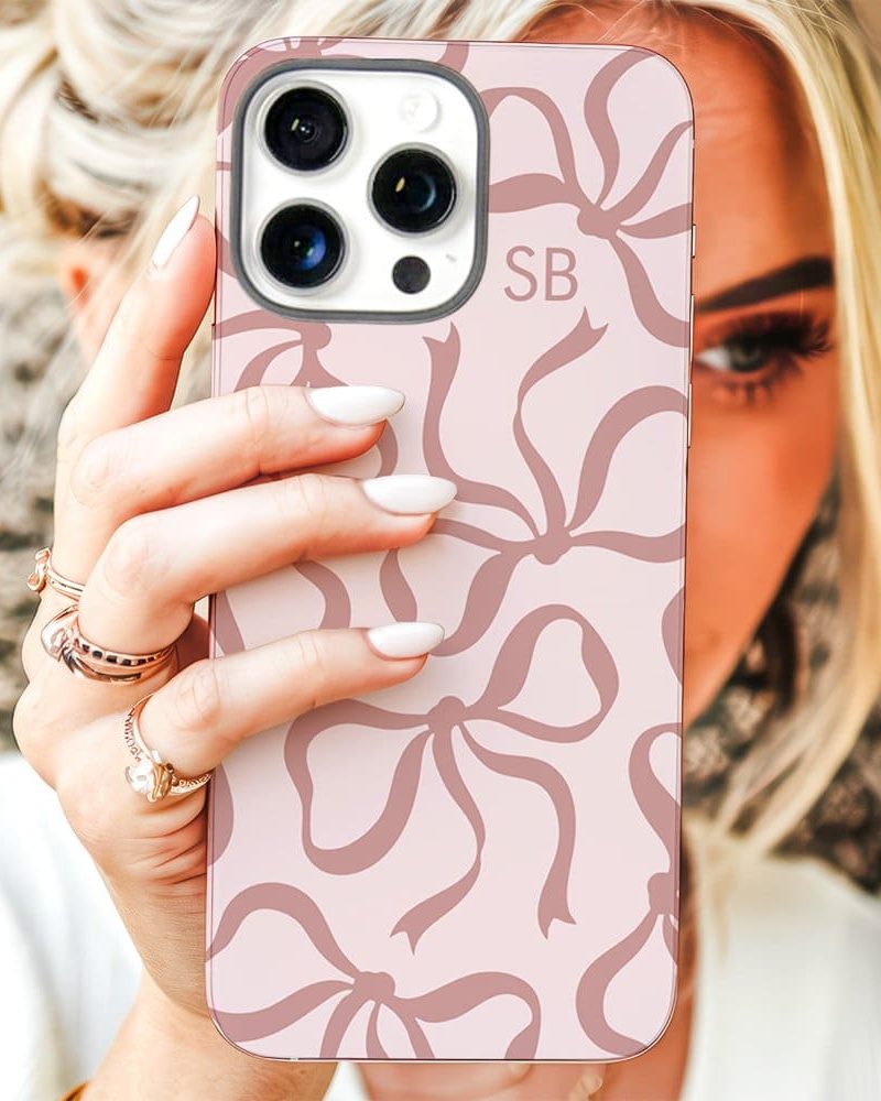 A woman holds up a pink iPhone case with a pattern of mauve bows. The initials SB are visible on the case.