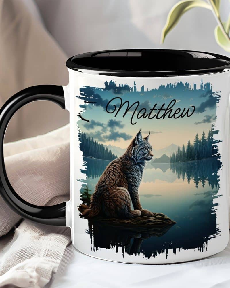 Black-handled mug with a lynx sitting by a lake at sunset; the name Matthew is printed on the mug.