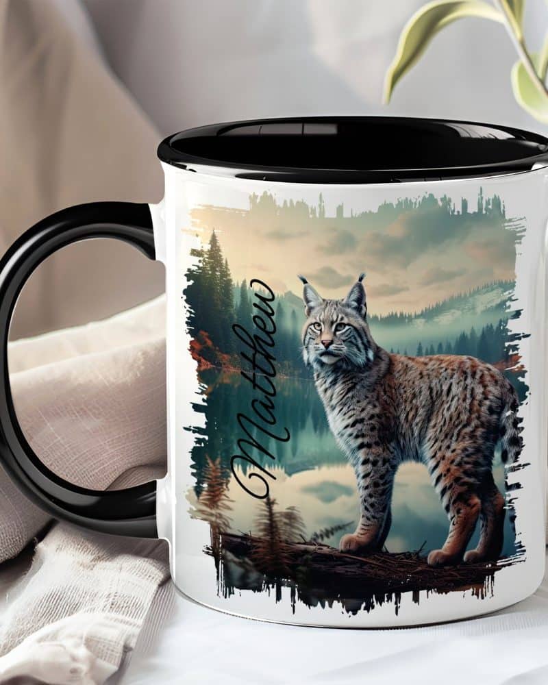 A black-handled mug features a lynx standing on a branch in a mountain landscape. The name Matthew is printed on the mug.