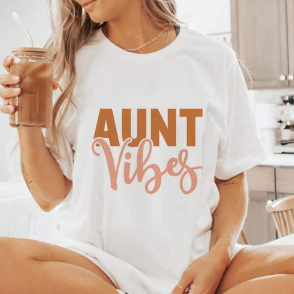 Woman Wearing A White T-Shirt That Says Aunt Vibes In Brown And Pink Lettering.