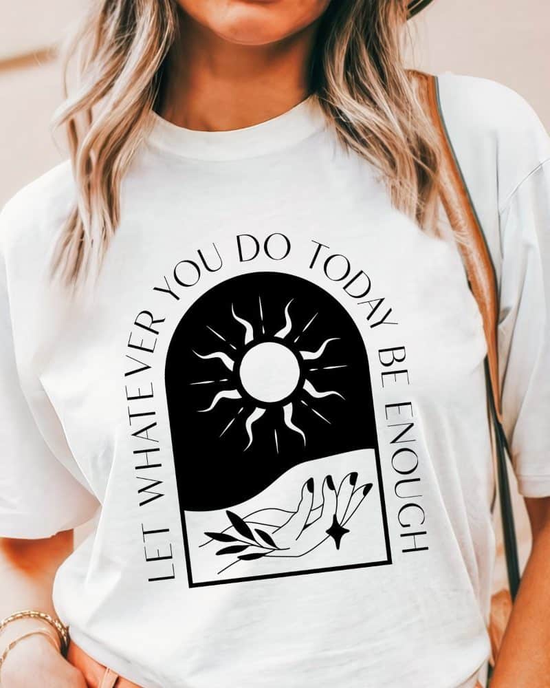 White t-shirt with a black arched graphic design. The design features a sun, a hand, and leaves, and the text Let Whatever You Do Today Be Enough.