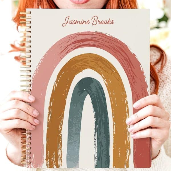 Person Holding Spiral-Bound Notebook Featuring A Pastel Rainbow Design. The Name Jasmine Brooks Is On The Cover.