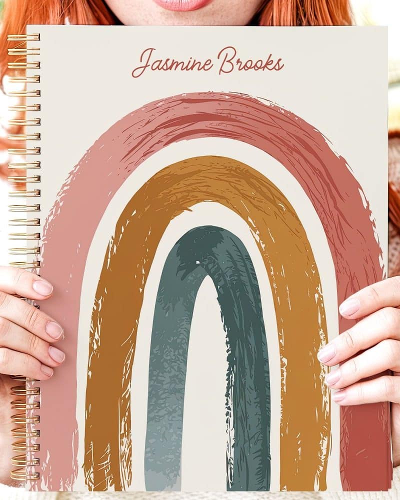Person holding spiral-bound notebook featuring a pastel rainbow design. The name Jasmine Brooks is on the cover.