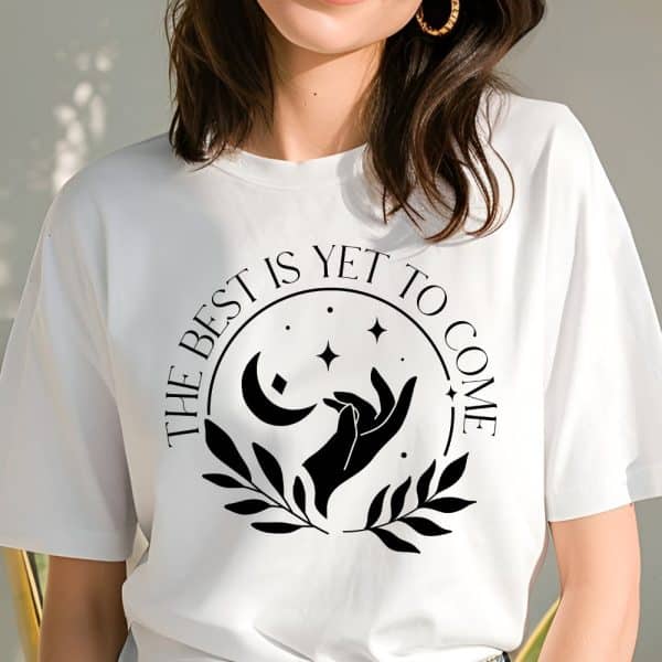 White T-Shirt With A Black Circular Design Featuring A Crescent Moon, Stars, Hands Reaching, And Leaves; Text Reads The Best Is Yet To Come.