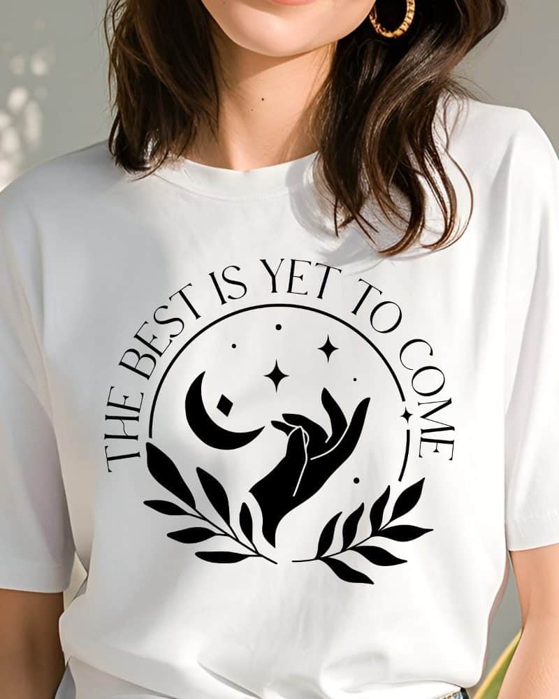 White t-shirt with a black circular design featuring a crescent moon, stars, hands reaching, and leaves; text reads The Best Is Yet To Come.