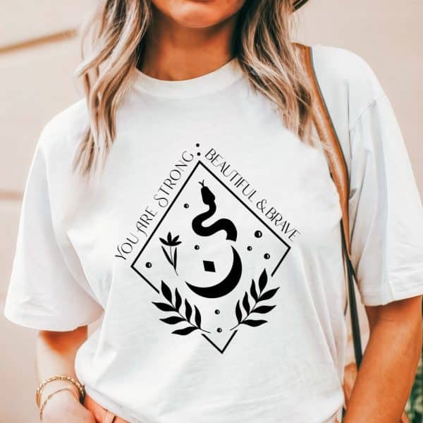 White T-Shirt With A Black Diamond-Shaped Design Featuring A Snake, Crescent Moon, Floral Elements, And The Text You Are Strong, Beautiful &Amp; Brave.
