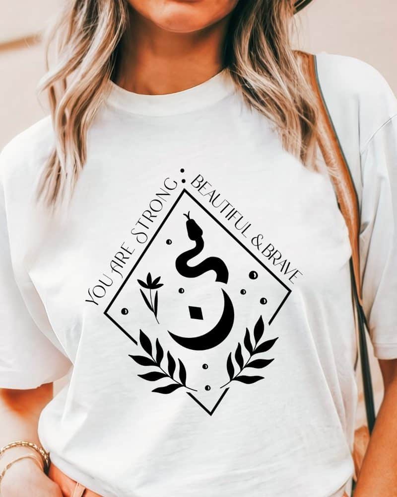 White t-shirt with a black diamond-shaped design featuring a snake, crescent moon, floral elements, and the text You Are Strong, Beautiful & Brave.