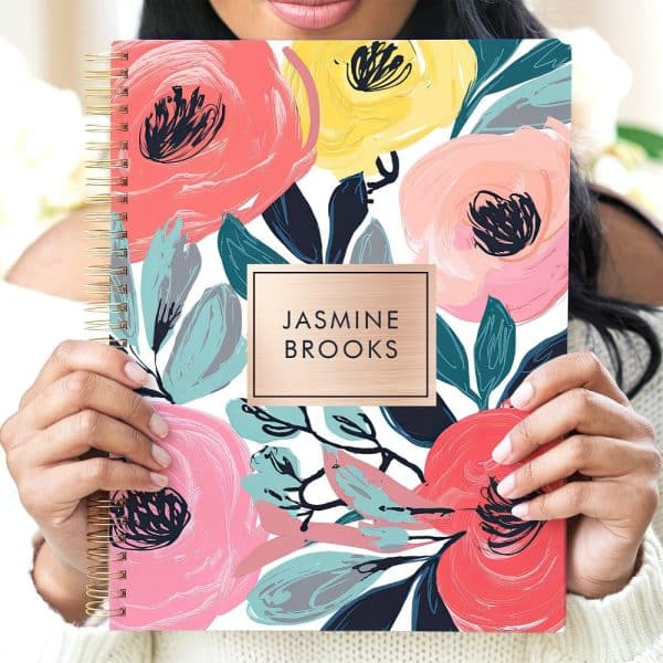 A Woman'S Hands Hold A Spiral-Bound Notebook With A Floral Design And The Name Jasmine Brooks.