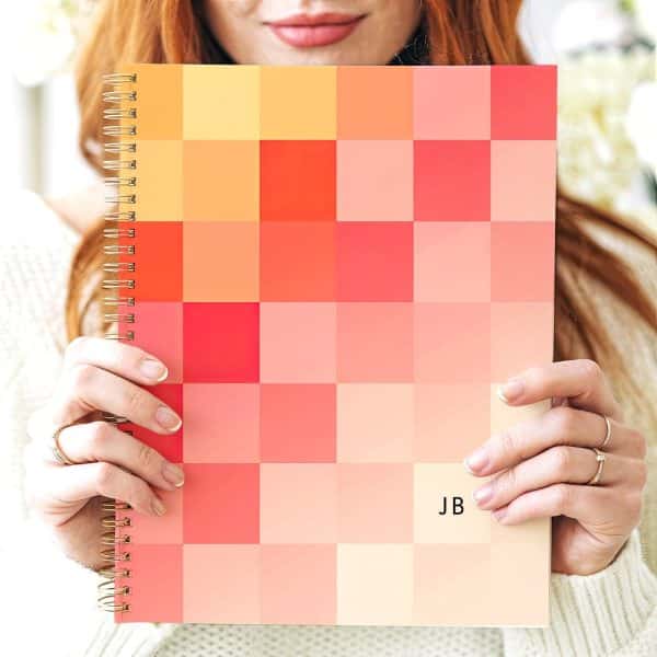 A Woman Holds A Spiral-Bound Notebook With A Pixelated Cover In Shades Of Orange And Pink. The Initials Jb Are Visible On The Cover.