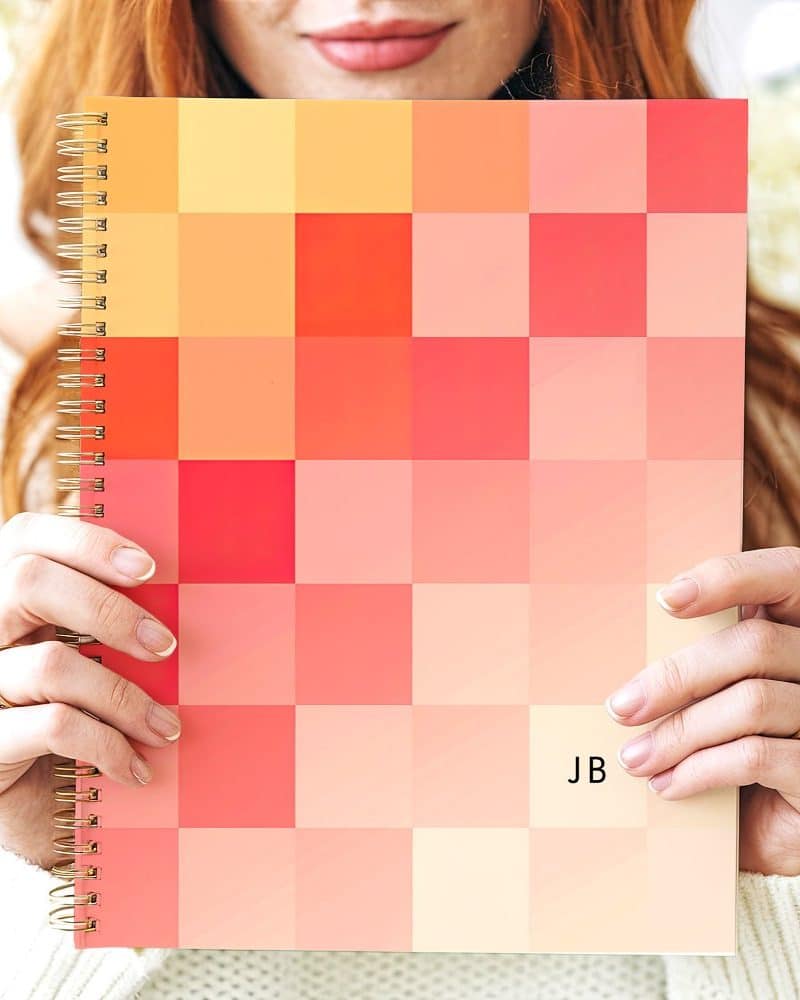 A woman holds a spiral-bound notebook with a pixelated cover in shades of orange and pink. The initials JB are visible on the cover.