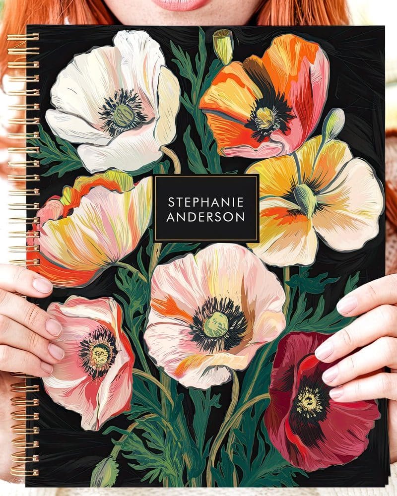 A woman holds a spiral-bound notebook featuring a dark background and a colorful floral design of poppies. The name Stephanie Anderson is printed on the cover.