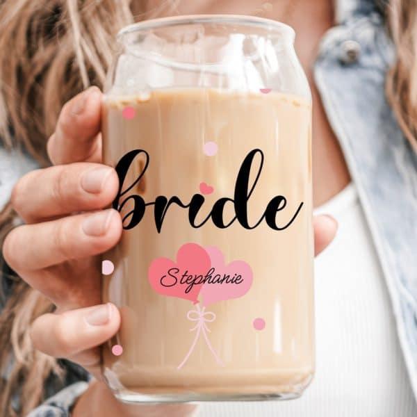 A Woman'S Hand Holds A Glass Iced Coffee Can With Bride And Stephanie Written On It, Along With Pink Heart Balloons.
