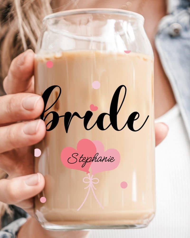 A woman's hand holds a glass iced coffee can with Bride and Stephanie written on it, along with pink heart balloons.