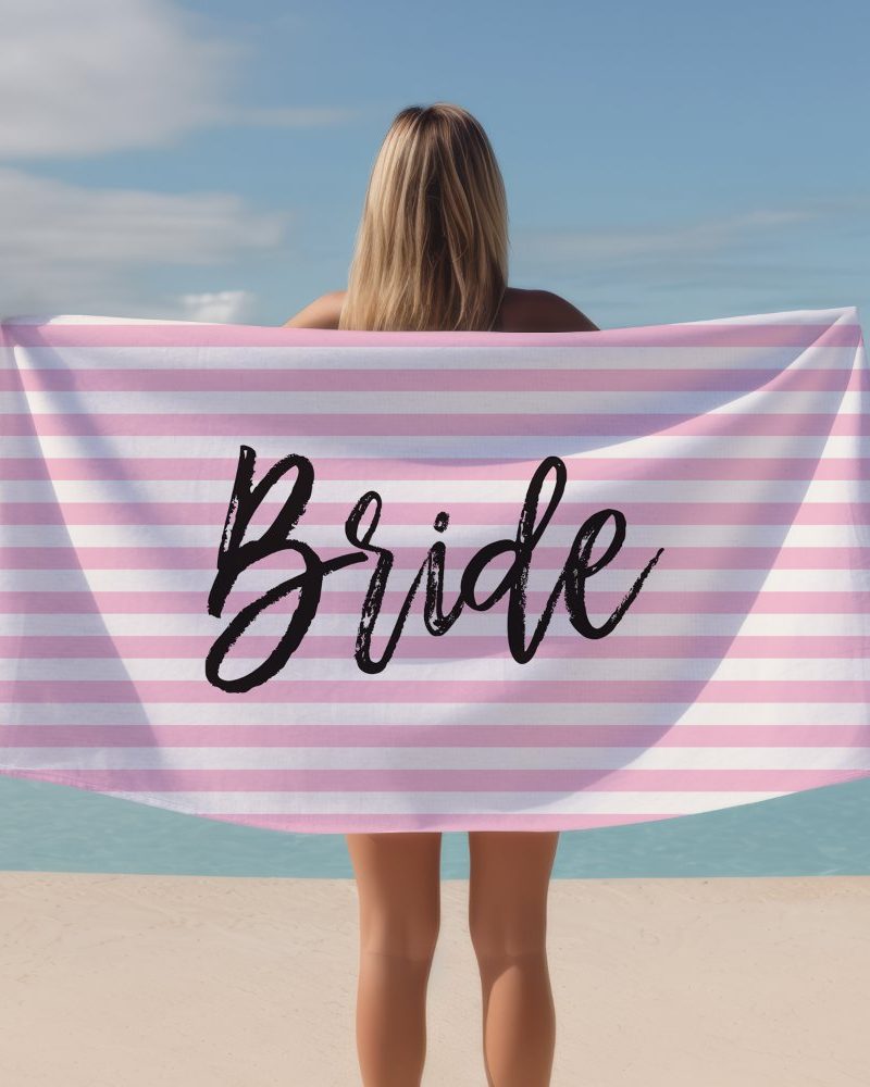 A woman holds a pink and white striped beach towel that says Bride in black lettering.