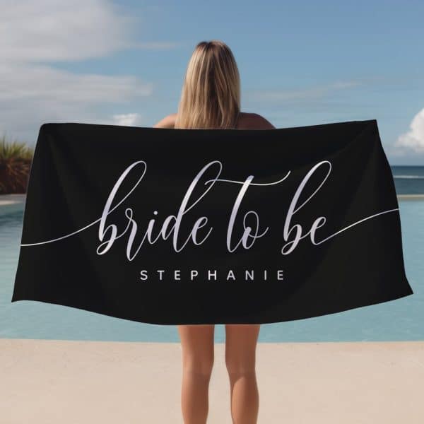 Woman Holding A Black Beach Towel That Says Bride To Be Stephanie In Elegant Script.
