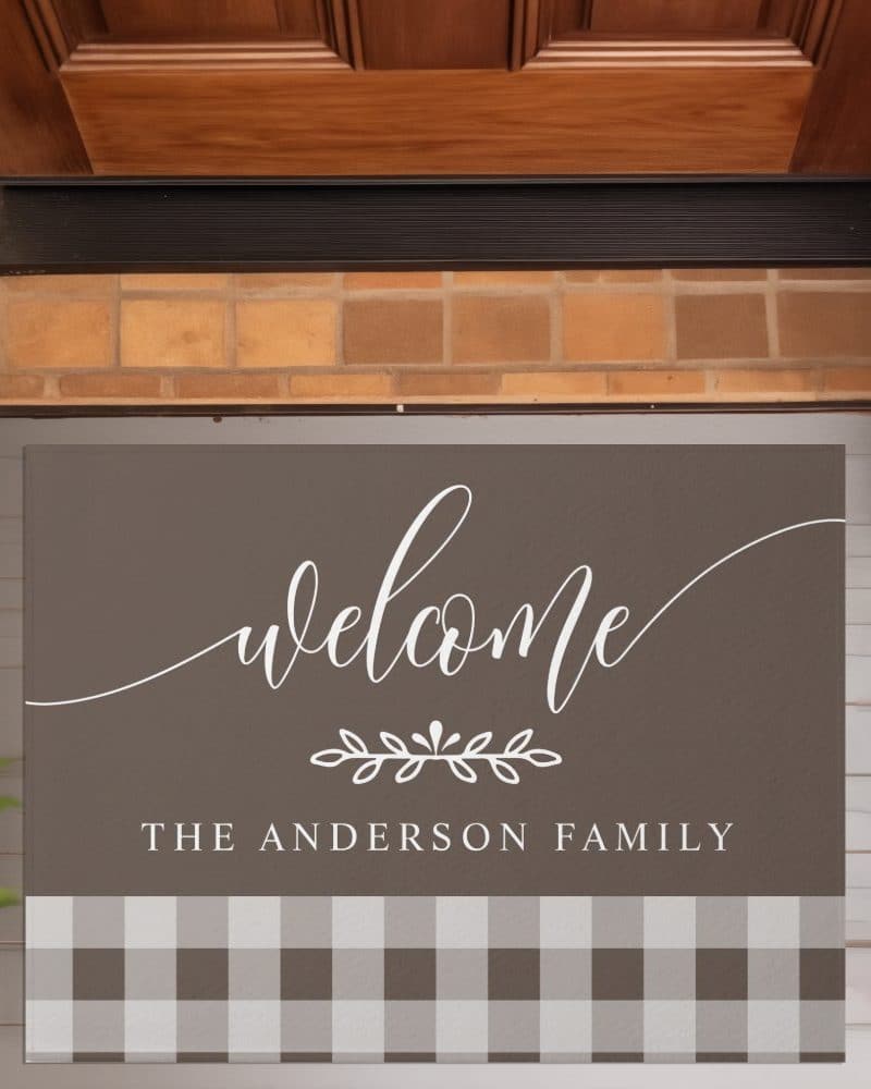 A welcome mat with the text Welcome The Anderson Family. The mat is brown with a white script font and a gingham bottom border.