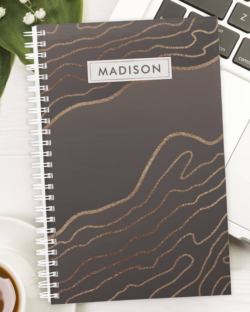 Spiral-bound notebook with dark gray cover and rose gold abstract lines; the name Madison is printed on the cover.