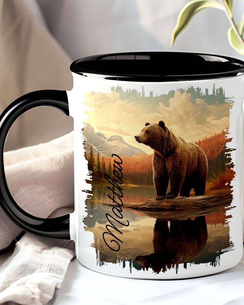 A black-handled mug with a grizzly bear image. The bear stands on a log by a lake, mountains in the background. The name Matthew is printed on the mug.