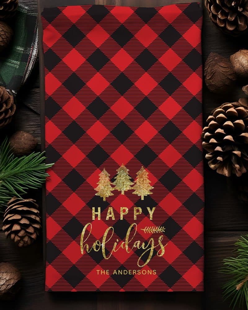 Red and black buffalo plaid kitchen towel with gold lettering that says Happy Holidays and The Andersons, and three gold trees.