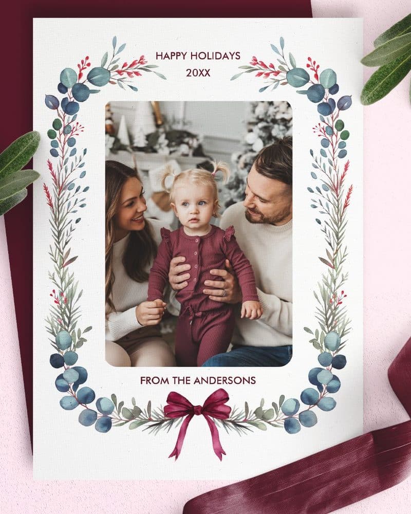 A holiday greeting card featuring a family photo; parents and young child. The photo is framed by a watercolor wreath.