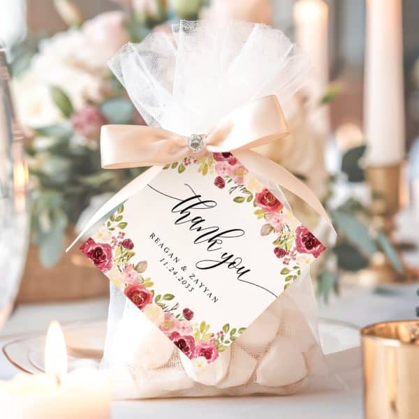 A Small White Favor Bag With A Pink Ribbon And A Floral Thank You Tag. The Tag Includes The Names Reagan &Amp; Zayyan And A Wedding Date.
