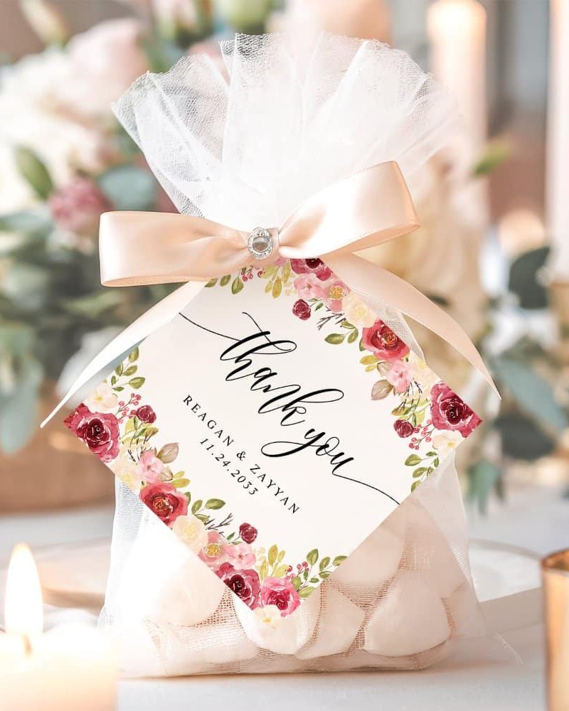 A small white favor bag with a pink ribbon and a floral thank you tag. The tag includes the names Reagan & Zayyan and a wedding date.