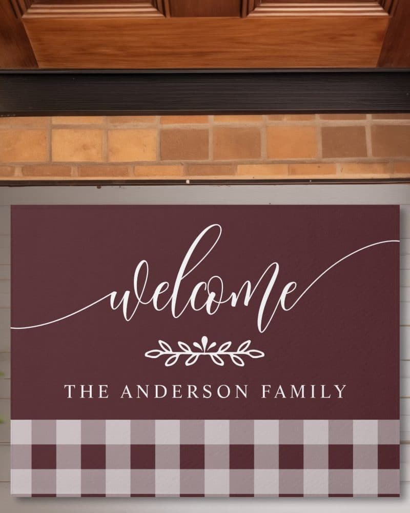 Burgundy welcome mat with white script lettering that says Welcome, a small floral design, and The Anderson Family. The bottom features a burgundy and white gingham pattern.