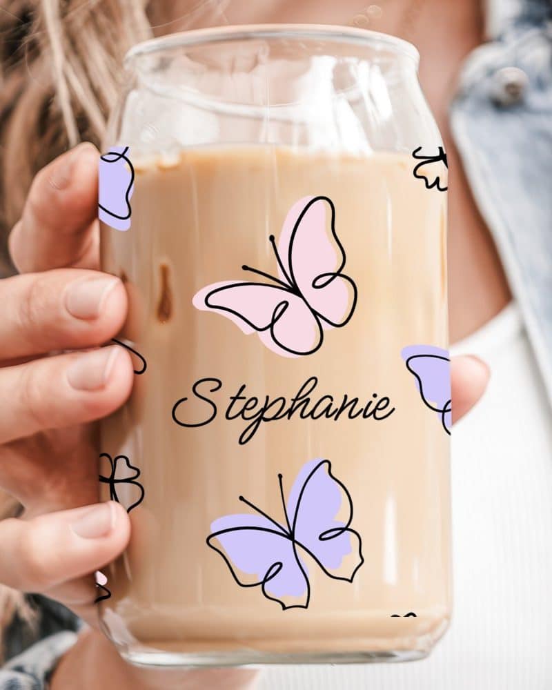 A hand holds a glass iced coffee cup decorated with butterflies and the name Stephanie.