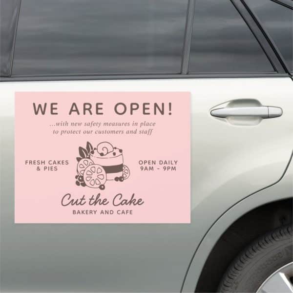 Pink Sign On A Car Announcing A Bakery And Cafe Is Open, Featuring A Cake Illustration And Daily Hours.