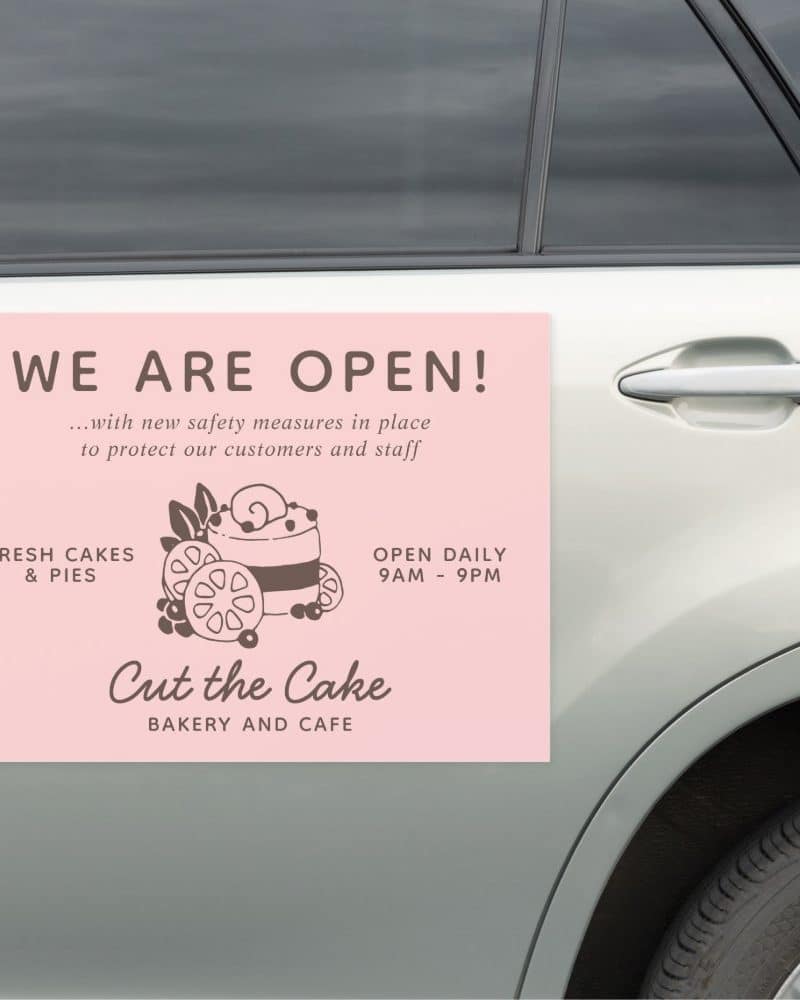 Pink sign on a car announcing a bakery and cafe is open, featuring a cake illustration and daily hours.