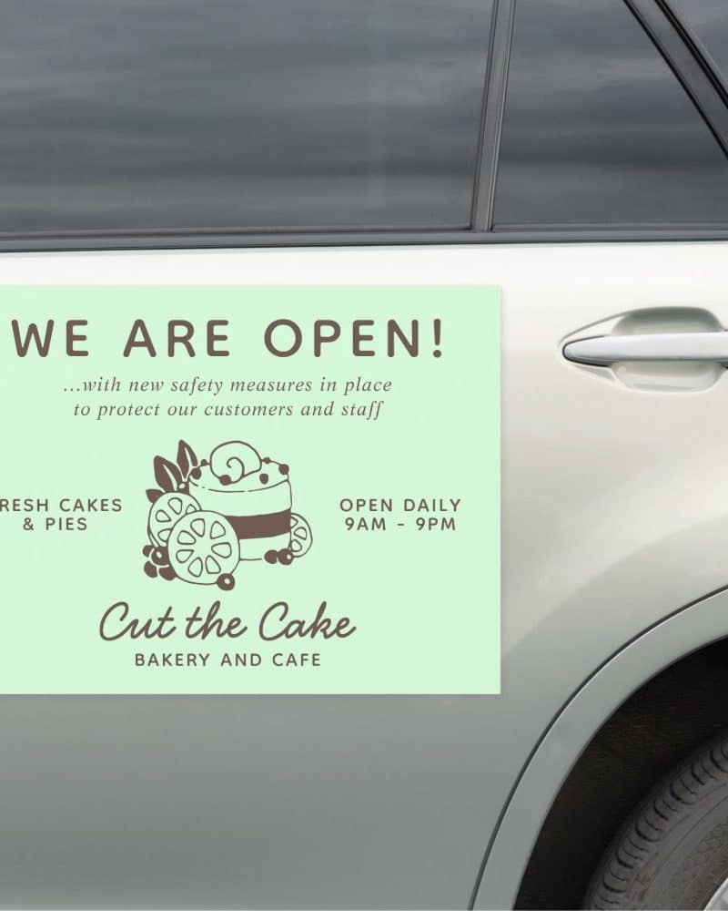 A light green sign on a car announces that Cut the Cake Bakery and Cafe is open, displaying hours and noting new safety measures are in place.