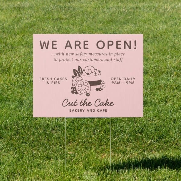 Pink Bakery Sign In Grass, Announcing They Are Open With Safety Measures In Place, Selling Cakes And Pies, Open 9 Am To 9 Pm Daily.