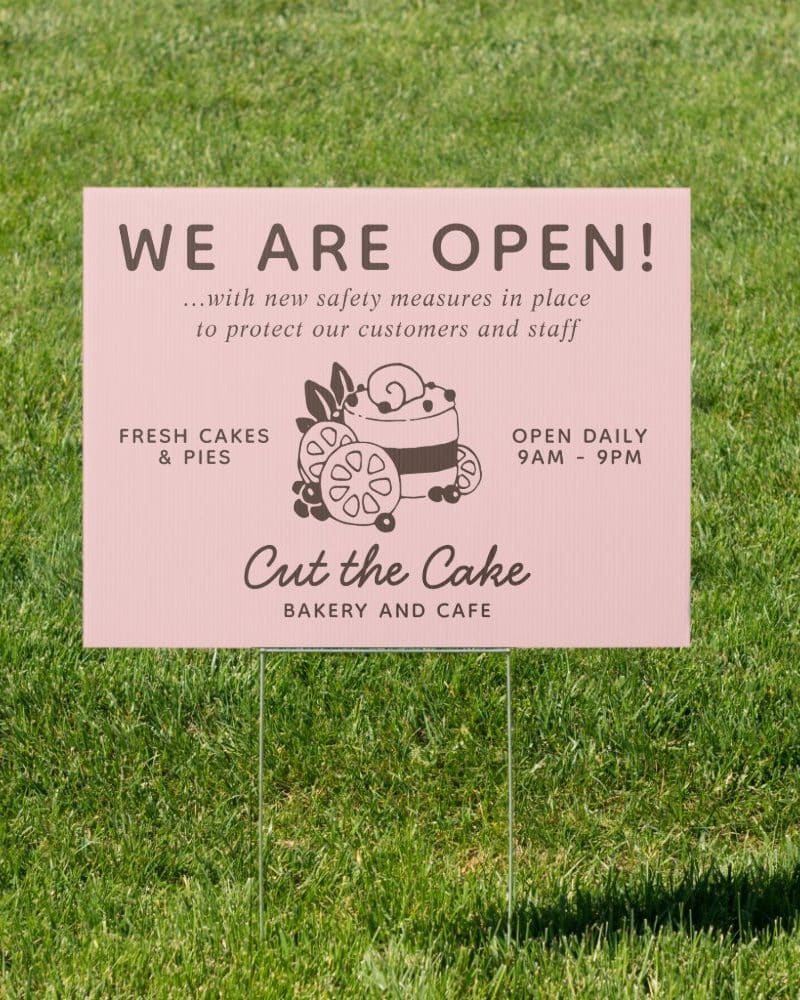 Pink bakery sign in grass, announcing they are open with safety measures in place, selling cakes and pies, open 9 AM to 9 PM daily.