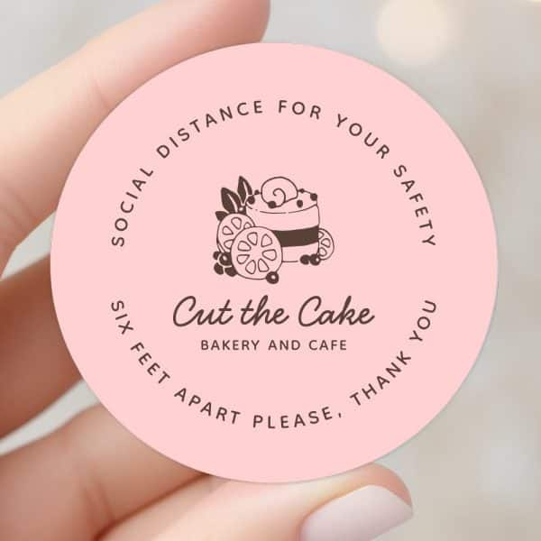 Pink Circle Sticker With A Logo For Cut The Cake Bakery And Cafe, Showing A Stylized Cake With Fruit. The Text Requests Social Distancing.