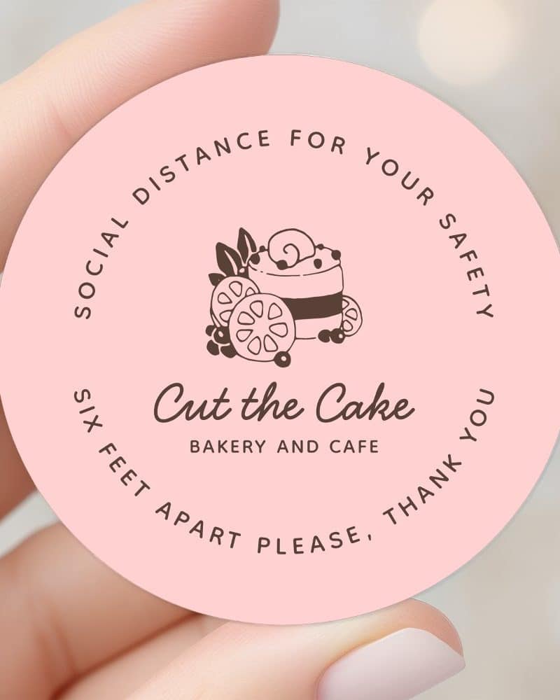 Pink circle sticker with a logo for Cut the Cake Bakery and Cafe, showing a stylized cake with fruit. The text requests social distancing.