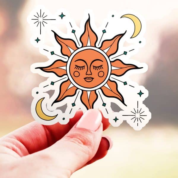Hand Holding A Sticker Of A Stylized Sun With A Face, Surrounded By Small Stars And Crescent Moons.