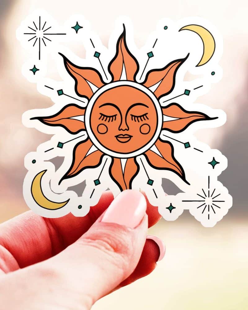 Hand holding a sticker of a stylized sun with a face, surrounded by small stars and crescent moons.
