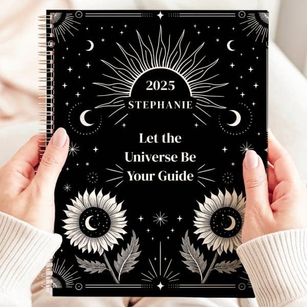 Two Hands Hold A 2025 Planner With A Black Cover. The Cover Features A Celestial Design With Suns, Moons, Stars, And Sunflowers In Cream. Text Reads Let The Universe Be Your Guide. The Name Stephanie Is Also Visible.